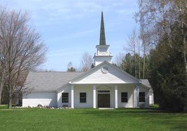 White church with a tall steeple in a green field under a clear blue sky.